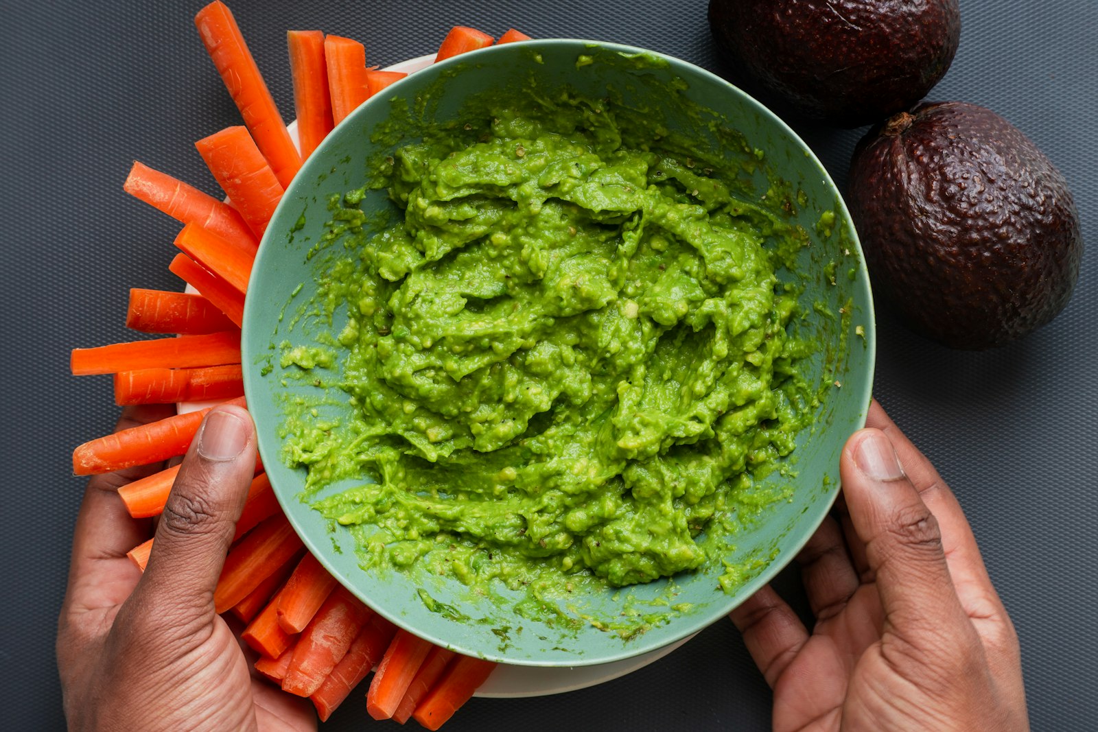 A bowl of green avocado and pea mash surrounded by fresh carrots